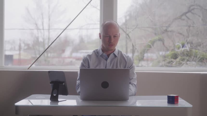 A bald man in a plaid shirt sits at a desk focused on a laptop with a tablet and a rubiks cube nearby he appears to be working or studying with a large window in the background offering a view of bare trees