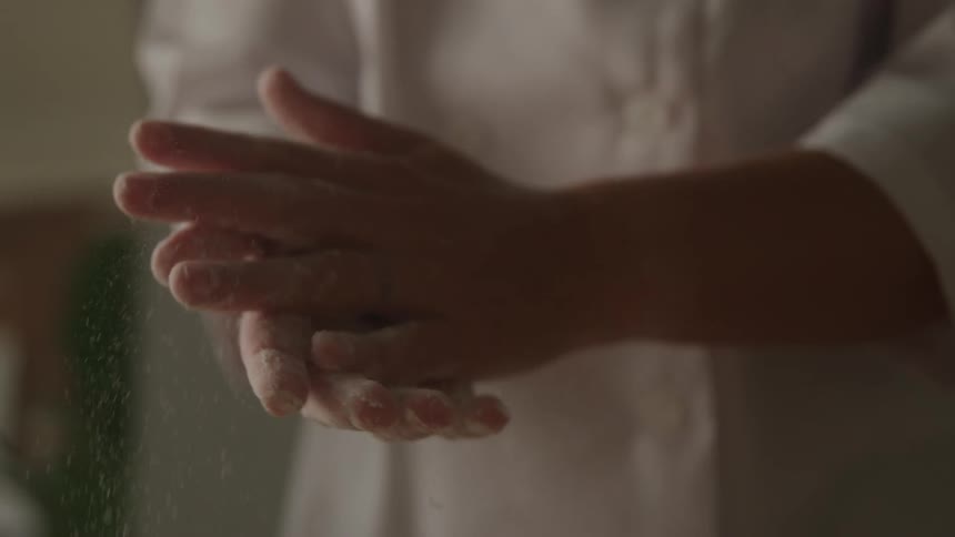 A person wearing a white chefs coat is rubbing their hands together creating a visible cloud of white powder likely flour the background is blurred focusing attention on the hands and the powder