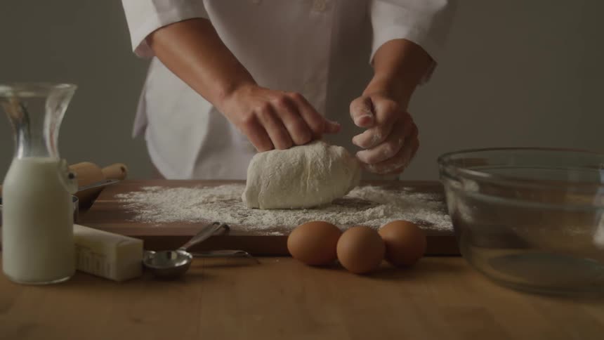 A chef kneads dough on a wooden board covered in flour with baking ingredients such as milk butter and eggs on the table beside a large glass bowl