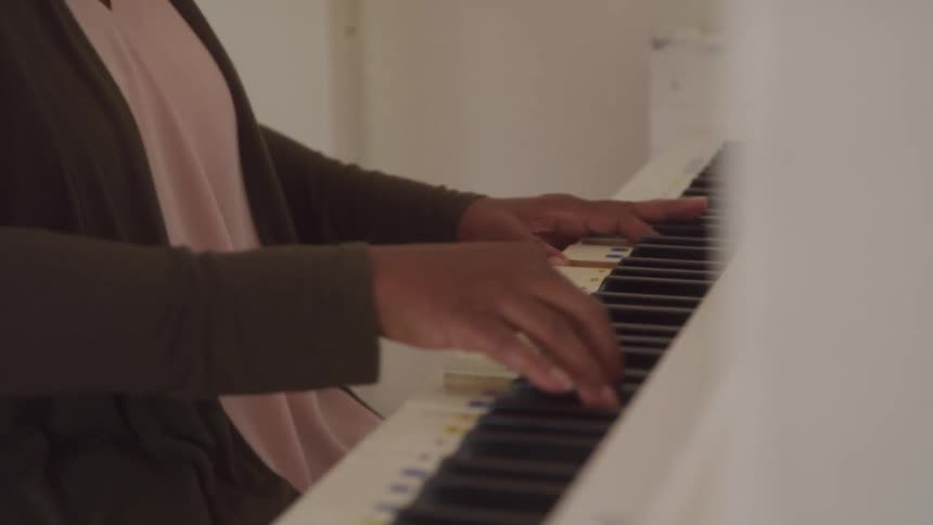 An older adult woman with her hair in a bun and wearing a green cardigan over a light pink top plays a white digital her hands moving across the keys looking down with a calm expression
