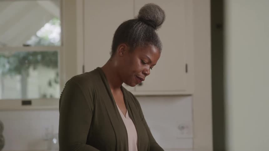 An older adult woman with her hair in a bun wearing a green cardigan and a light colored top stands in a kitchen chopping colorful apples on a cutting board with apples on the counter and white cabinets and a window in the background
