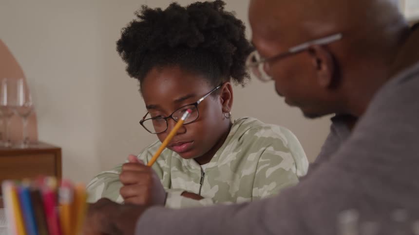 An adult man likely an instructor wearing glasses and a grey shirt sits at a wooden table helping a young female student with curly hair and glasses as she writes in an open book