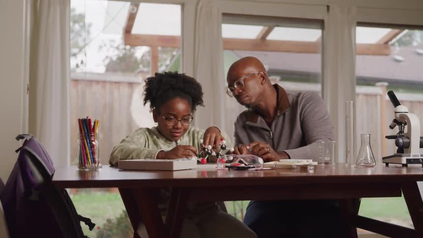 A young female student with curly hair and glasses works on a molecular model at a wooden table while an adult male likely an instructor leans in to assist her with a microscope and beakers on the table and a window looking out at a backyard