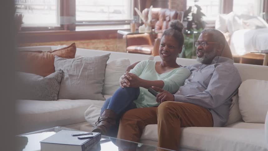 An older adult couple sits comfortably on a light colored sofa in a living room with the man holding a television remote as they both watch television and converse surrounded by decorative pillows and furniture