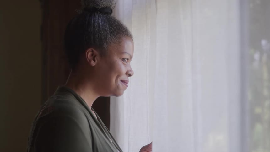 A side profile shot captures a woman with grey hair tied in a bun wearing a green longsleeved shirt as she smiles while looking out of a window the bright window with white vertical blinds contrasts with the darker indoor setting