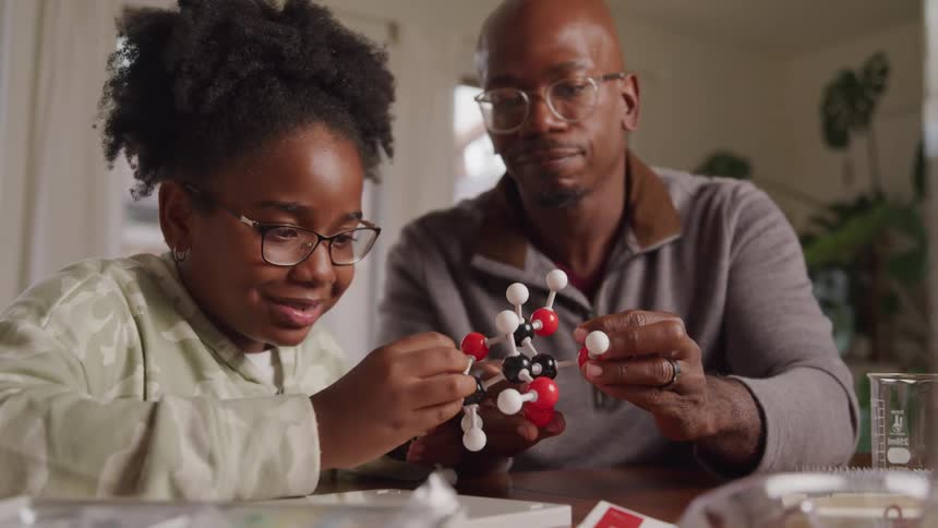 A father and his daughter sit at the dining table wearing glasses having fun learning together as they examine a molecular model