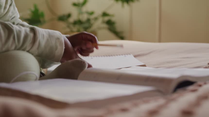 A child wearing light colored socks and pants sits on a bed with an open book in the foreground holding a pencil and writing on a notebook doing homework