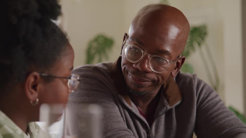 A bald man wearing glasses and a grey jacket is intently focused on a task holding a pencil while a young woman in glasses is partially visible next to him also looking down at what they are working on they appear to be studying or collaborating
