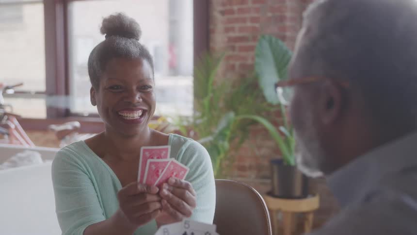 An elderly man with gray hair plays a card game with a woman indoors at midday both smiling
