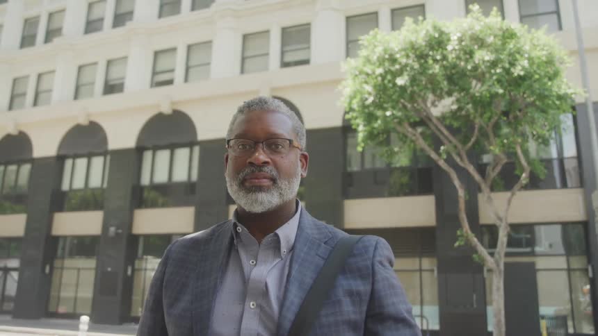 A professionally dressed black man stands near a city street appearing thoughtful as sunlight filters through the trees and buildings