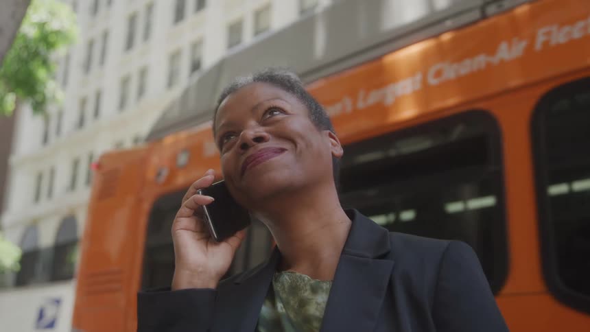 An older woman with short grey hair wearing a dark blazer and a patterned top holds a phone to her ear and smiles broadly while looking upwards with an orange bus and a city building in the background