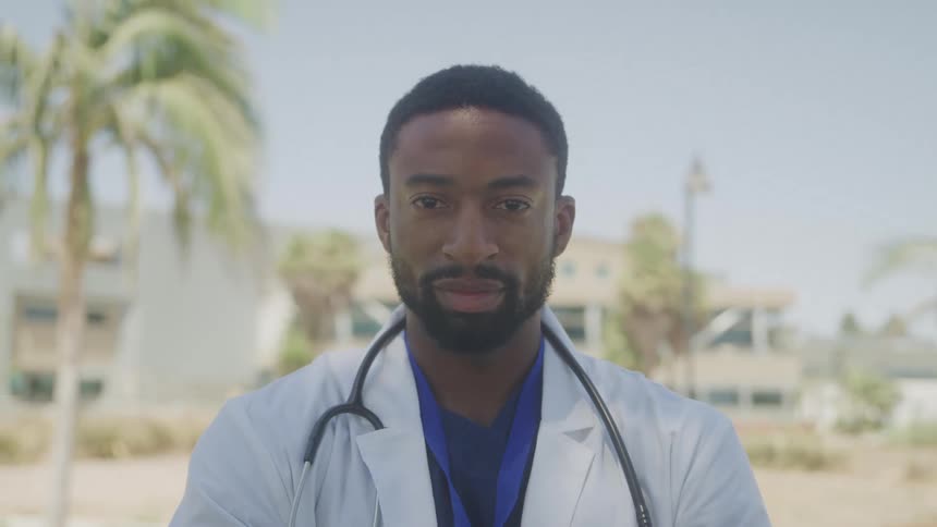 A doctor glaring meanly in a white coat with a stethoscope looks calmly ahead standing outdoors near palm trees