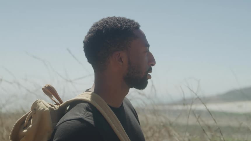 A man with a beard and short dark hair is shown in profile looking towards the right side of the frame with a thoughtful or distant expression he is wearing a dark tshirt and a lightcolored backpack suggesting he might be outdoors or on a hike with