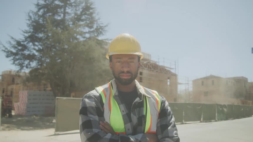 A construction worker wearing a hard hat and safety vest stands confidently with arms crossed at an active building site