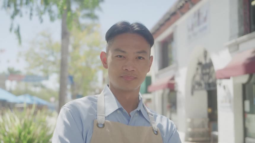 A man with short dark hair wearing a light blue collared shirt and a light colored apron looks directly at the camera with a subtle smile standing outdoors on a street lined with buildings and foliage