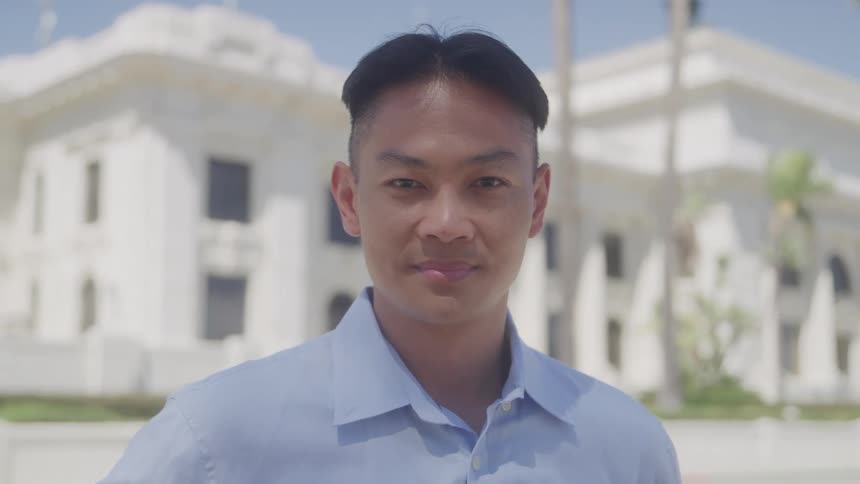 A young adult man with short dark hair wearing a light blue collared shirt looks directly at the camera with a subtle smile standing outdoors in front of a light colored building