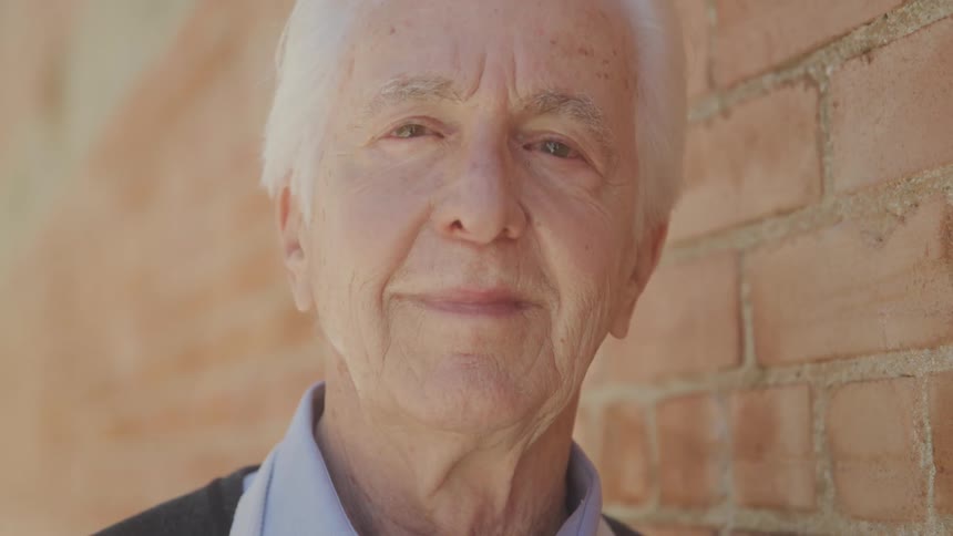 An elderly man with gray hair wearing an apron rests against a brick wall at midday