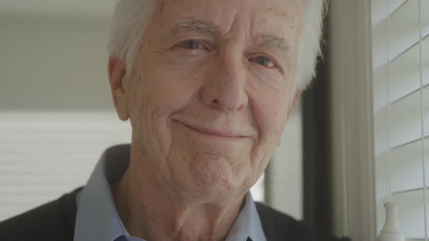 A greyhaired man with visible wrinkles around his eyes and mouth smiles while looking out a window with horizontal blinds the scene suggests a serene introspective moment as he closes and then reopens his eyes
