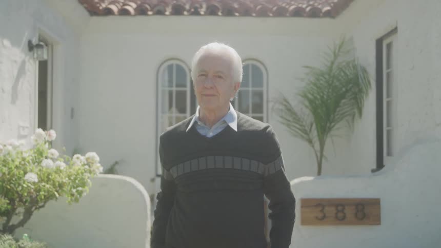 An elderly man with white hair wearing a striped sweater and a light blue collared shirt is seen standing outdoors in front of a white building with arched windows and the number 388 visible beside the door he initially looks away and then turns to smi