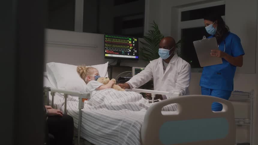 A young child with a blue face mask lies in a hospital bed holding a teddy bear as two medical professionals attend to her while her mother sits by the bedside in the room