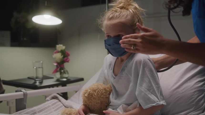 A young girl with blond hair wearing a blue face mask and holding a teddy bear sits up in a hospital bed as a female nurse uses a stethoscope to listen to her back with flowers and a glass of water on a bedside table