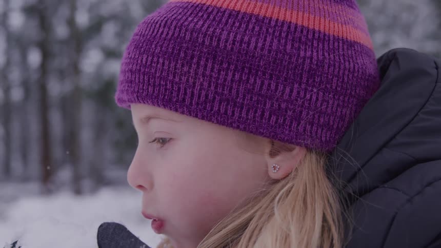 A young girl with long blond hair wearing a purple and orange knitted hat and dark coat blows powdered snow from her gloved hands with trees in a snowy background