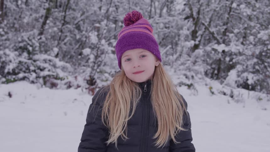Out in the woods at midday a young girl with blonde hair stands in the snow wearing a winter jacket and hat