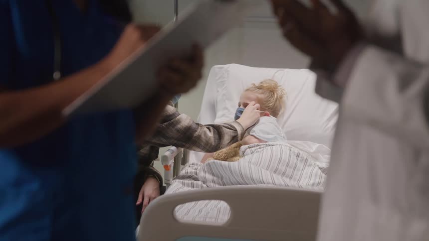 Two doctors talk in the foreground and in the background a mother with her young daughter lying in a hospital bed
