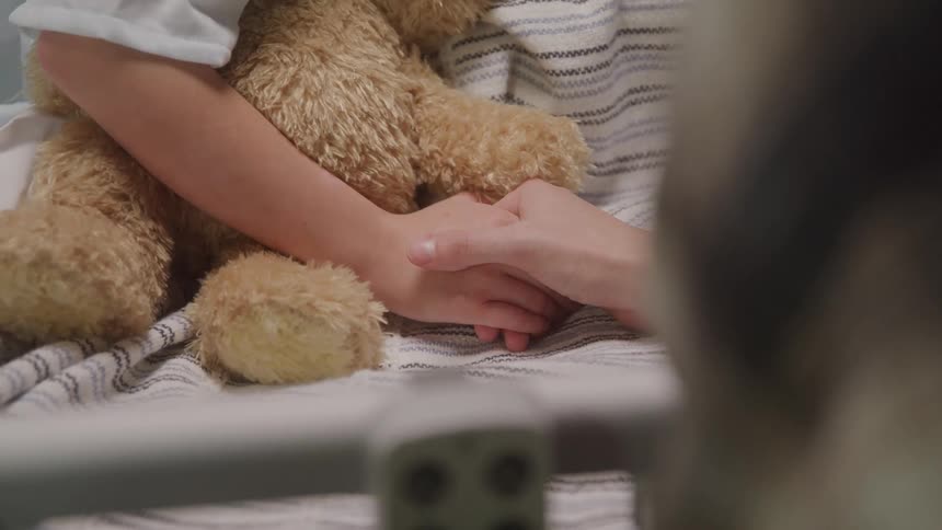 A close view shows a young childs hands resting on a white striped blanket holding a brown teddy bear while another persons hand reaches out to gently clasp the childs hand