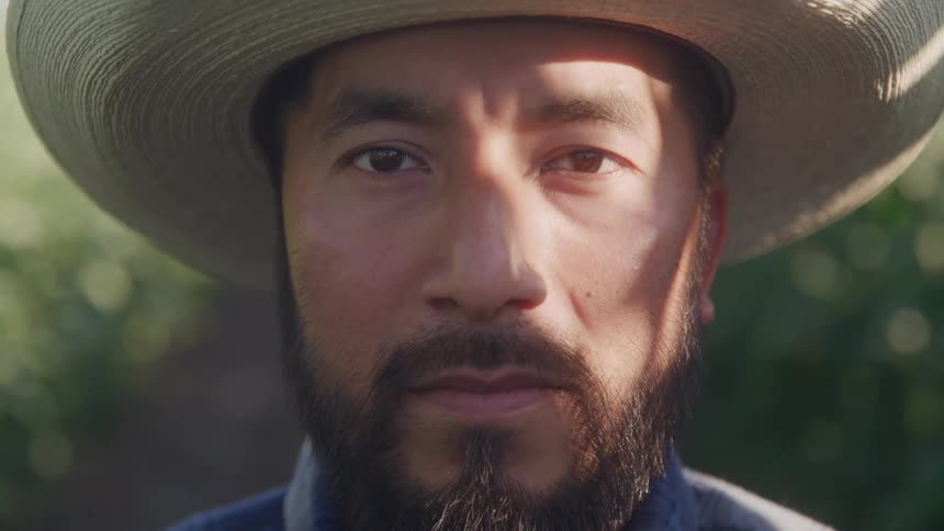 The face of a bearded man wearing a cowboy hat staring straight ahead while his face is partially illuminated by natural light