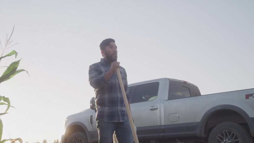 Man next to a pickup truck in a field weeding and then taking off his hat