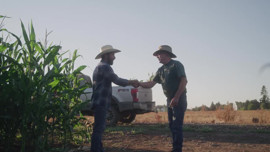 Two men wearing sun hats stand in a cornfield on a sunny day shaking hands and smiling with a truck parked nearby