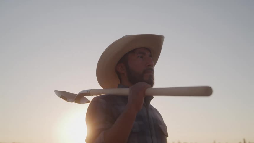 At sunset a man stands outdoors wearing a sunhat and holding a shovel over his shoulder