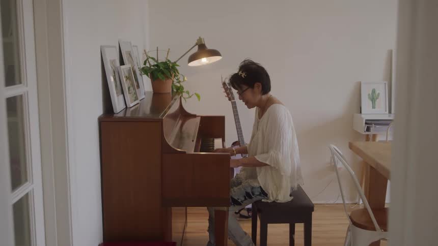 A woman with dark hair and glasses wearing a light colored top and patterned pants sits on a bench playing a brown upright piano with a guitar leaning against it and a lamp plants and framed pictures