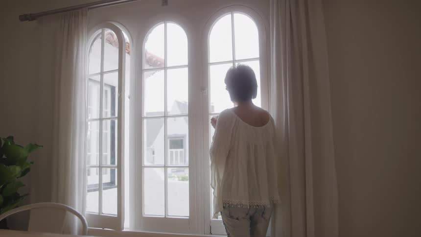 A woman stands with her back to the camera looking out a large arched window with sheer white curtains while a plant and part of a chair are visible in the foreground