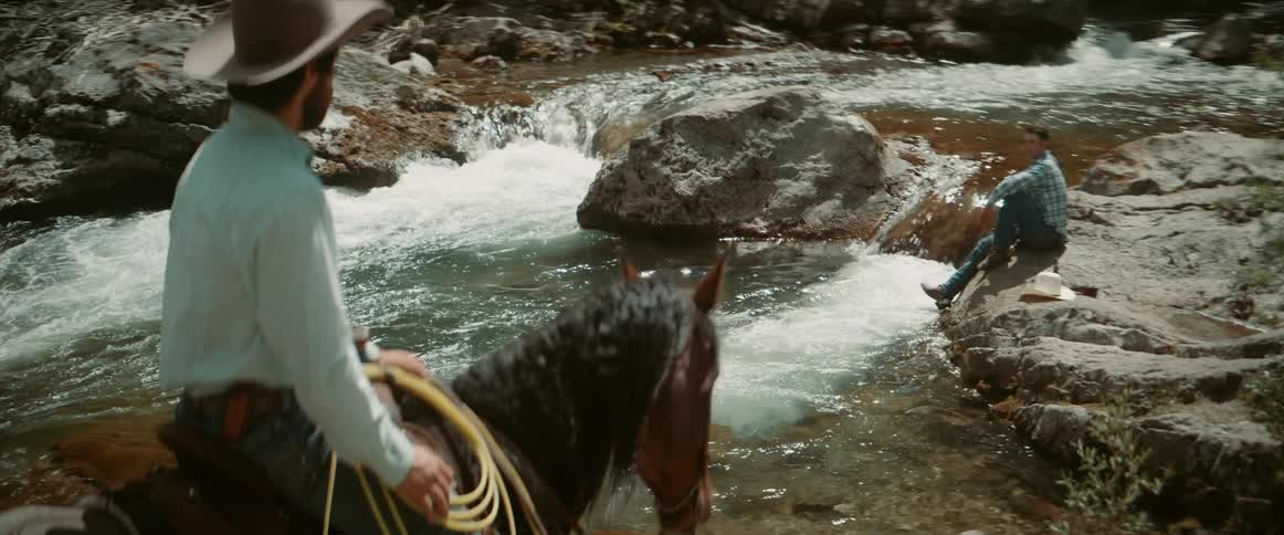 Horse rider pauses to talk to a man sitting on the riverbank drinking