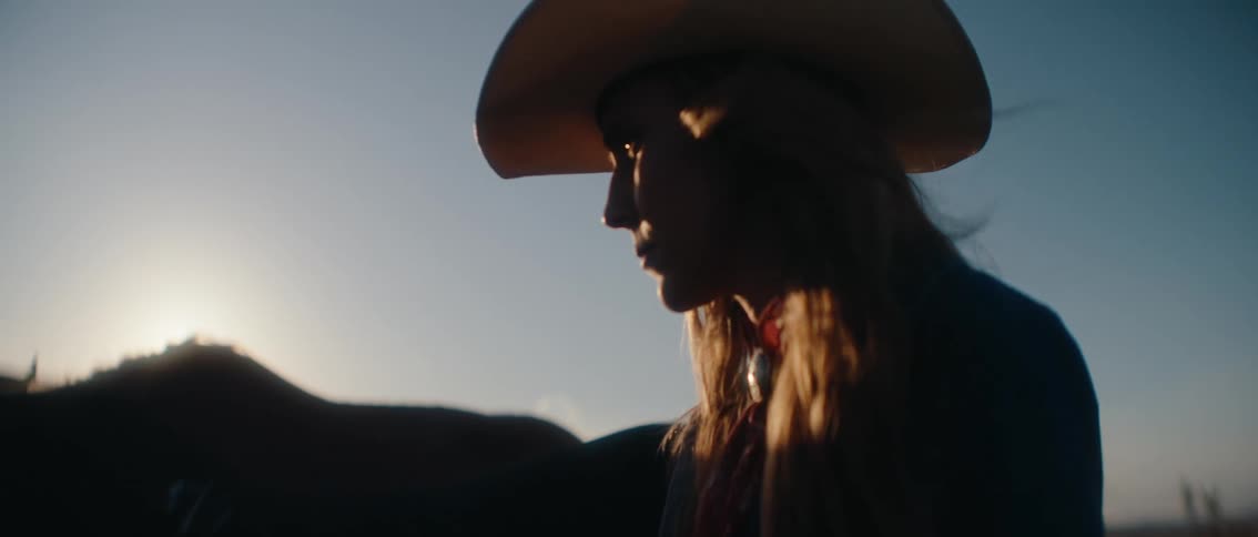 A woman wearing a cowboy hat walks beside a horse inside a ranch at sunset