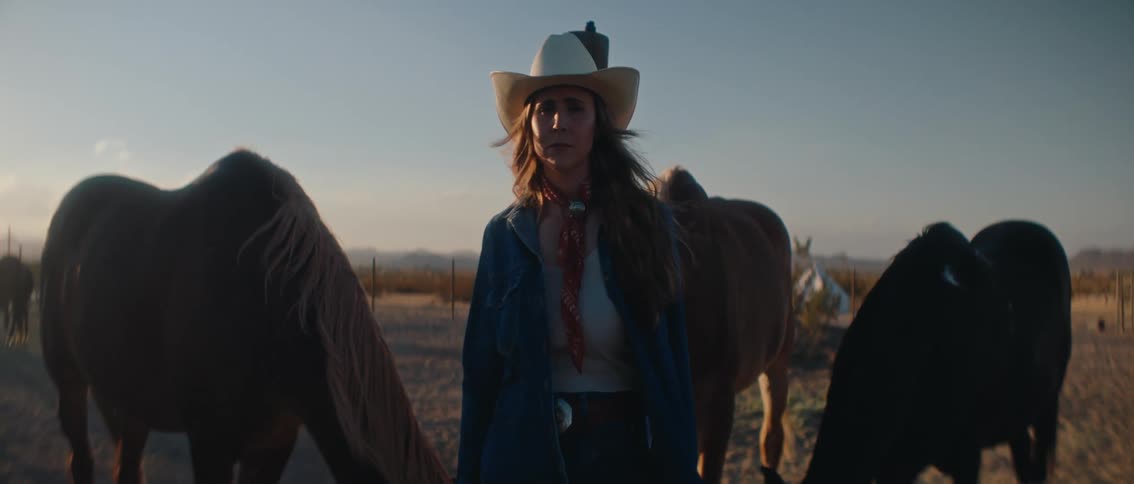 A person in a cowboy hat and denim jacket stands looking towards the right with horses partially visible around them in a dusty outdoor setting the scene is bathed in the warm light of what appears to be late afternoon or early morning with mountains