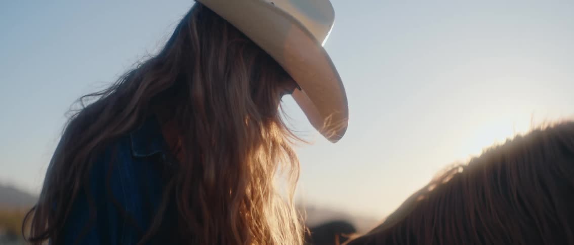 A woman with long light colored hair wearing a cowboy hat and a blue jacket stands near a horse and a bright sky in the background during sunset