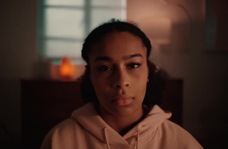 A young woman is looking directly at the camera with a neutral expression on her face the room is dimly lit with a blurry lamp visible in the background
