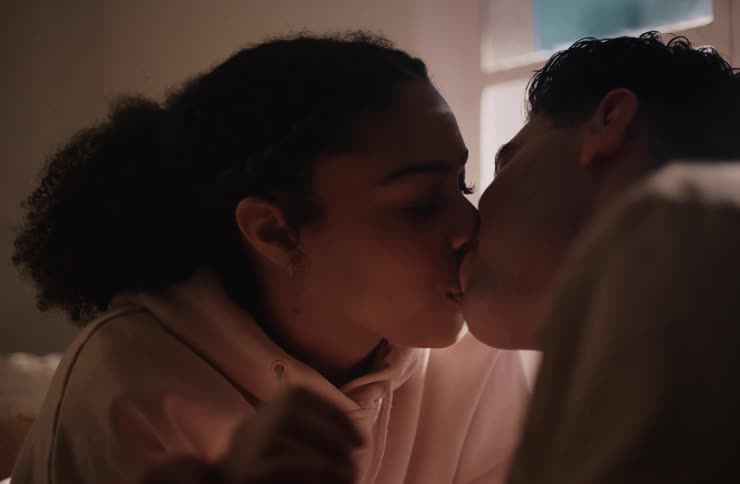 A young adult couple leans in close to each other a woman with dark curly hair in a light colored hoodie looking at a man and then they kiss and pull apart both smiling subtly