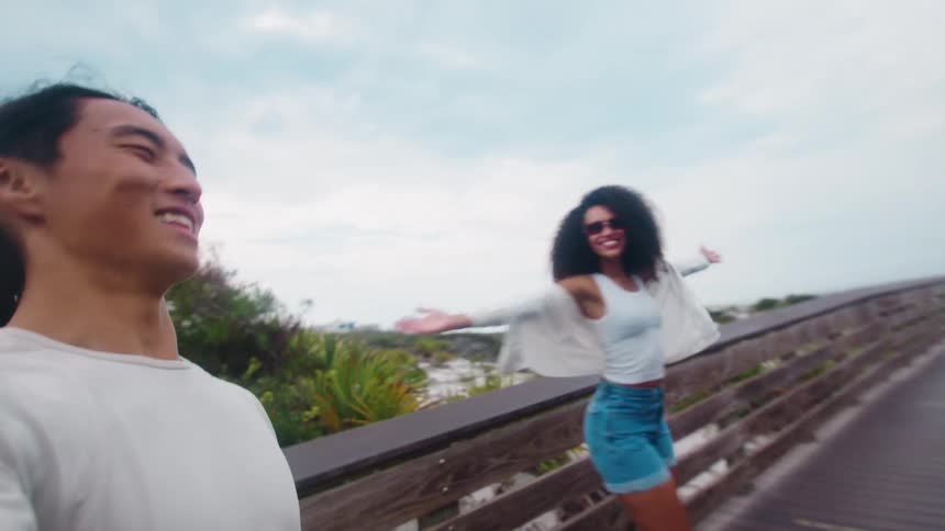 A smiling couple is walking together on a wooden boardwalk the woman is wearing sunglasses and has her arms stretched out to the sides while the man is holding a camera taking a video selfie of them