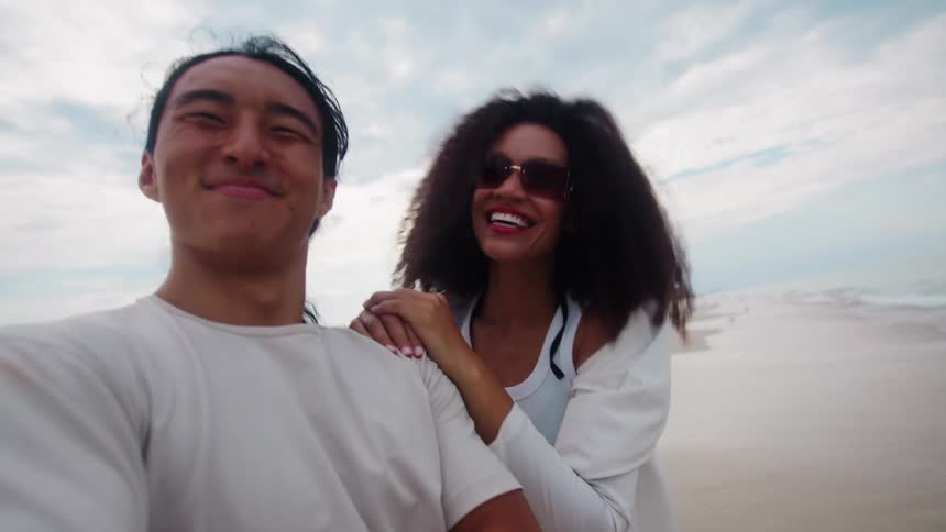 A man and a woman who appear to be a couple take a selfie video on a beach the man in a light shirt holding the camera with a wide smile as the woman with curly hair and sunglasses wearing a white top stands beside him smiling and raising her arm