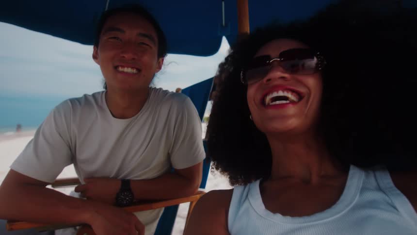 A man and a woman are sitting together on a beach under a blue umbrella the woman wearing sunglasses is smiling widely at the camera while the man sits next to her with a smile on his face