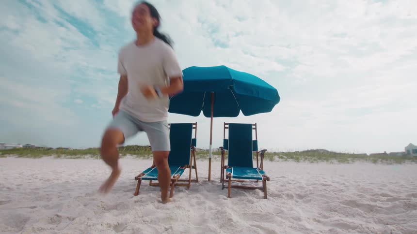 A couple sitting under an umbrella on the beach suddenly get up as the woman starts running on the sand and the man runs after her barefoot at midday
