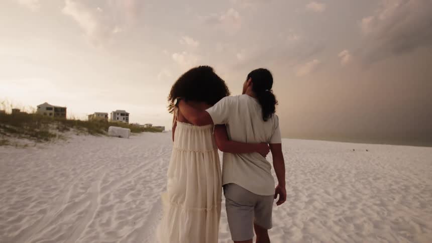 A couple walks down the beach with their arms around each other shown from behind