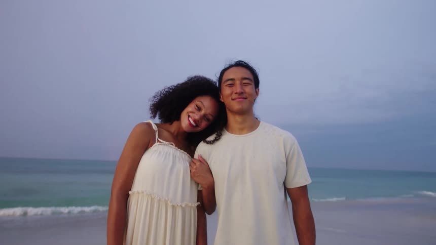 A wide shot of a smiling couple standing on a beach with the woman resting her head on the mans shoulder as they both look at the camera