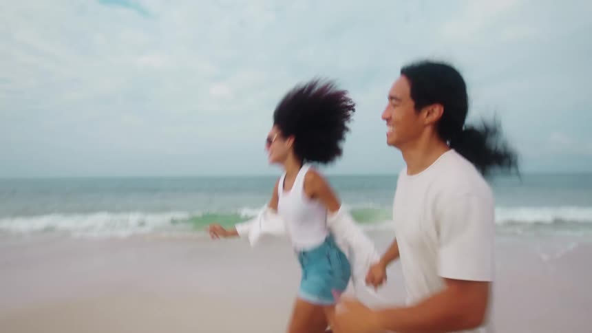 A smiling couple is running joyfully on a beach the woman is leading and the man is following holding her hand as they run toward the camera