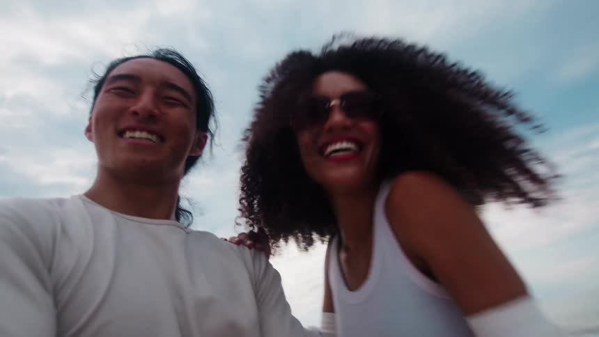 A couple having a fun time on the beach both smiling and laughing as they walk around with the woman wearing sunglasses at midday