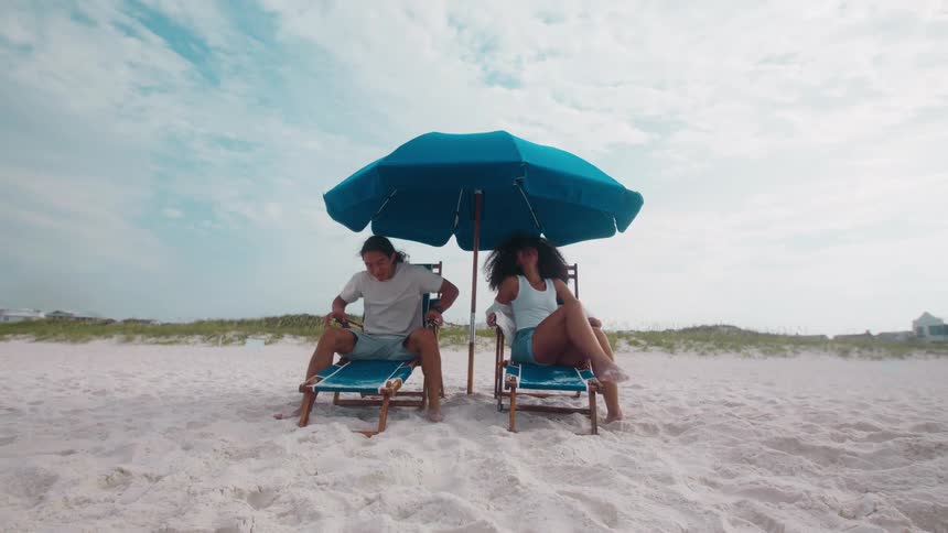 Two people relax under a beach umbrella on lounge chairs at the shore